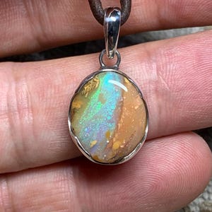 May include: An oval-shaped pendant featuring a polished opal gemstone with iridescent flashes of green, blue, and gold set in a smooth silver bezel. The pendant is attached to a dark brown leather cord, showcasing the natural colour variations.
