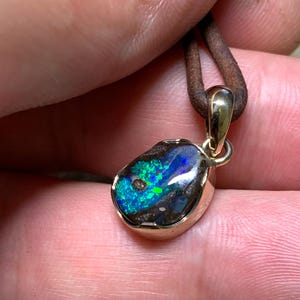 May include: A gold pendant featuring an opal stone with blue and green hues. The opal is set in a gold bezel and suspended from a brown leather cord.