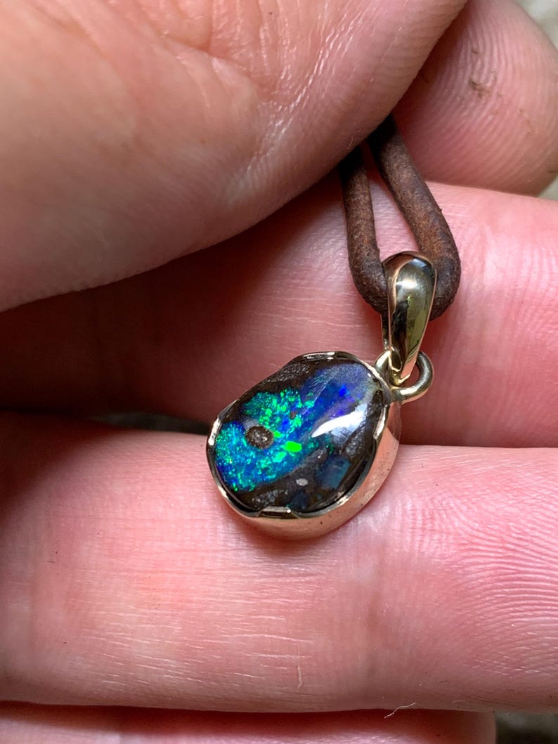 May include: A gold pendant featuring an opal stone with blue and green hues. The opal is set in a gold bezel and suspended from a brown leather cord.