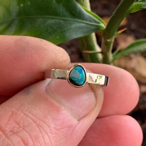 May include: A gold-toned ring with a teardrop-shaped opal gemstone. The opal has a dark blue and green color with a gold bezel setting. The ring band is a simple, polished design.