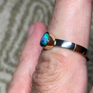 May include: A gold and silver ring featuring a teardrop-shaped opal gemstone with vibrant blue and green hues. The ring has a polished gold bezel setting and a silver band. The opal's iridescent colors are prominent.