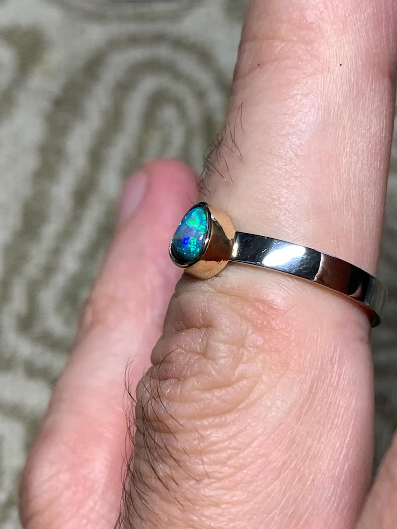 May include: A gold and silver ring featuring a teardrop-shaped opal gemstone with vibrant blue and green hues. The ring has a polished gold bezel setting and a silver band. The opal's iridescent colors are prominent.