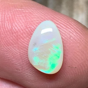 May include: A teardrop-shaped opal gemstone, polished to a high sheen. The opal displays a milky white base with vibrant flashes of green and blue. The stone is smooth and appears to be held by a finger.
