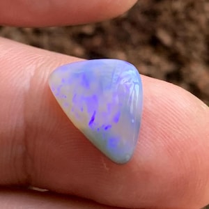 May include: A polished opal gemstone, showing a mix of colours. The stone has a triangular shape with a light blue base, and vibrant purple and yellow flashes. The opal is held between fingers.
