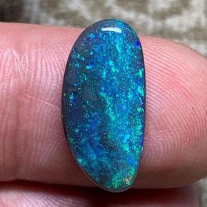 May include: An oval-shaped opal gemstone, polished to a smooth finish. The stone exhibits a striking play-of-colour, with shimmering blues, greens, and teals against a dark background. The opal is held between fingers, highlighting its natural beauty.