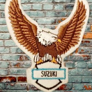 May include: A white embroidered patch with a brown eagle with outstretched wings. The eagle is standing on a blue and white shield with the word "SUZUKI" written on it.