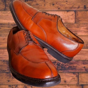 May include: A pair of brown leather lace-up shoes with a pointed toe. The shoes have a leather sole and a stitched welt. The shoes are on a wooden surface.