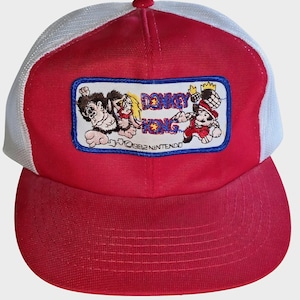May include: Red and white trucker hat featuring a Donkey Kong patch. The patch displays Donkey Kong, Pauline, and Mario characters with the text "DONKEY KONG" and "©1982 NINTENDO". The hat has a red brim and crown with a white mesh back.