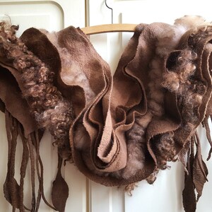 Può includere: Una sciarpa di lana feltrata marrone con un design a balze e frange lunghe. La sciarpa è appesa a un appendiabiti di legno.