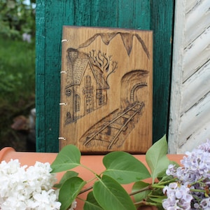 Può includere: Un diario in legno con un design intagliato raffigurante una casa, una montagna, un tunnel e binari ferroviari. Il diario è rilegato con spago bianco e viene esposto con fiori bianchi e viola.