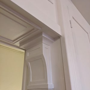 May include: A white door frame with decorative moulding. The frame features a curved bracket, rectangular panels, and a detailed top section. The door and frame are painted white, creating a clean and classic appearance. The design suggests a traditional style.