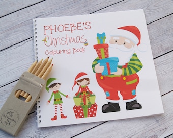 Puede incluir: Un libro para colorear blanco con encuadernación en espiral. La portada presenta un Papá Noel de dibujos animados sosteniendo regalos, dos duendes y el texto "PHOEBE'S Christmas Colouring Book".