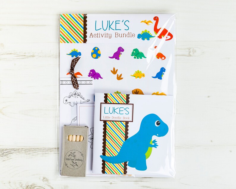 May include: A dinosaur-themed activity bundle for kids named "LUKE'S Activity Bundle". The bundle includes a coloring book, a sticker sheet, and a set of crayons. The coloring book features a blue dinosaur with a green leaf in its mouth. The sticker sheet has a variety of colorful dinosaurs and footprints. The crayons are brown and have a label that says "Little Twigs Stationery".