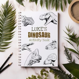 Op de afbeelding: Een zwart-wit activiteitenboek met de titel "LUKE'S DINOSAUR Activity Book". De omslag toont illustraties van verschillende dinosaurussen, waaronder een Tyrannosaurus Rex, een Triceratops en een Stegosaurus.