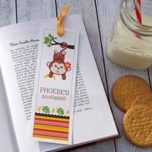 May include: A colourful bookmark featuring a cartoon monkey hanging from a tree branch with a banana. The bookmark has a striped border and the text "PHOEBE'S BOOKMARK" in black letters.