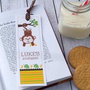 May include: A colourful bookmark featuring a cartoon monkey hanging from a tree branch with a banana. The bookmark has a yellow, orange, and green striped background with a green leaf design. The text "LUKE'S BOOKMARK" is printed in brown letters.