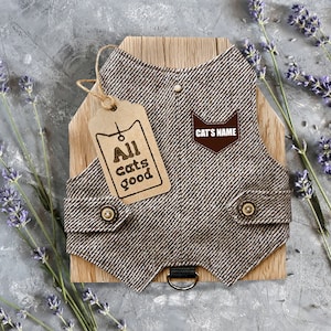Pode incluir: Um arnês para gato de tweed castanho e branco com um remendo de couro castanho que diz "CAT'S NAME". O arnês tem um botão castanho e um anel em D preto. Uma etiqueta de papel castanho com o texto "All cats good" está presa ao arnês.