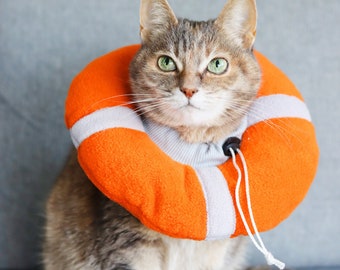 May include: A tabby cat wearing an orange and white life preserver-style protective collar. The collar is made of soft, plush fabric and has a white strap that ties at the back of the neck.