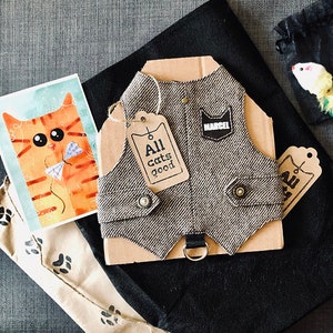 Puede incluir: Un chaleco de tweed gris con una etiqueta negra que dice "MARCEL" y una etiqueta marrón que dice "All cats good". El chaleco está sobre un fondo marrón y negro.