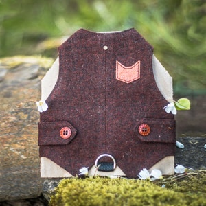 Può includere: Un gilet di feltro marrone con una piccola tasca in pelle arancione chiaro sul davanti. Il gilet ha due bottoni marroni e un anello metallico sul fondo.