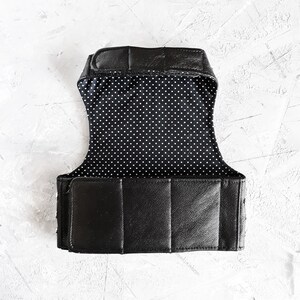 K&ouml;nnte beinhalten: Schwarze Lederweste mit wei&szlig;em Punktmusterfutter. Die Weste hat verstellbare Tr&auml;ger und einen Schnallenverschluss.