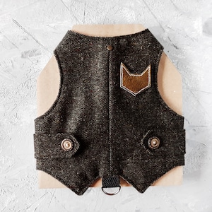Op de afbeelding: Een bruine tweed hondenvest met een leren vos patch en twee knoopsluitingen. De vest heeft een zwarte band met een metalen ring.