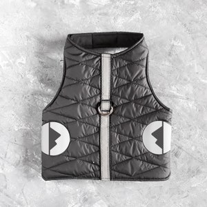 Puede incluir: Un arnés acolchado gris para perros con una franja reflectante central y una anilla en D metálica para sujetar la correa. El arnés presenta dos logotipos circulares blancos y grises en los laterales. Diseñado para la comodidad y la seguridad.