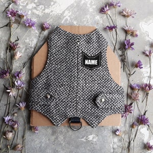 Può includere: Un'imbracatura per cani in tweed grigio e bianco con una toppa nera che dice "NAME".