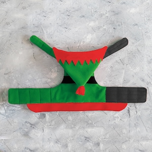 May include: A green and red elf costume for a pet. The costume features a green pointed hat with a red zig-zag trim, a red collar, and a green body with black accents. The costume has adjustable black straps.