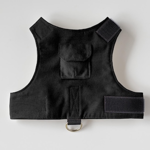 Pode incluir: Colete preto para animais de estimação com superfície texturizada, com um pequeno bolso e fechos de velcro. Inclui um anel em D de metal na parte inferior. O colete é projetado para animais de estimação e é feito de um material durável.