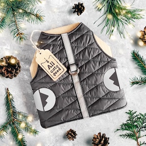 May include: A black quilted dog vest with reflective accents and a white mountain logo. The vest has a brown tag attached that says "All cats good".