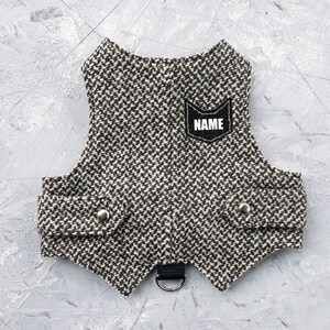 Puede incluir: Un arnés para perro de tweed marrón y blanco con un parche negro que dice "NAME". El arnés tiene un anillo en D negro para sujetar una correa.