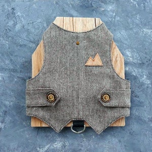 Puede incluir: Un chaleco para perro de color marrón y negro con estampado de espiga, con un parche de montaña de cuero marrón y dos botones dorados. El chaleco tiene una correa negra con un anillo en D plateado.