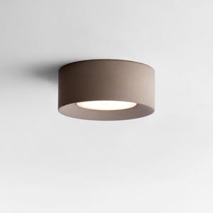 Könnte beinhalten: Eine moderne, zylindrische Deckenleuchte. Die Leuchte hat eine neutrale, taupefarbene Außenseite und eine helle, eingelassene Lichtquelle. Die Leuchte ist für die bündige Montage an der Decke konzipiert und bietet eine saubere, minimalistische Ästhetik.
