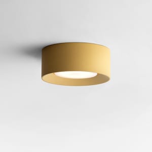 Könnte beinhalten: Eine senfgelbe, zylindrische Deckenleuchte mit einer hellen, weißen Lichtquelle. Die Leuchte ist bündig an einer weißen Decke montiert und wirft einen weichen Schatten. Das Design ist minimalistisch und modern.