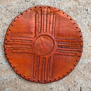 May include: A brown leather round wall hanging with a stitched design of a Native American symbol, the Zia symbol, in the center.