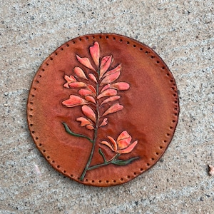 May include: A round leather coaster with a stitched edge features a tooled design of a pink flower with green leaves.
