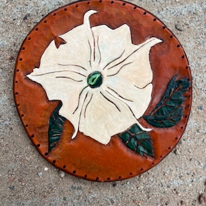 May include: A round leather coaster with a white flower design and green leaves. The coaster has a brown background and is stitched around the edge.