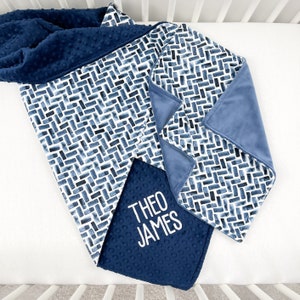 K&ouml;nnte beinhalten: Eine marineblaue Babydecke mit einem geometrischen Aquarellmuster in Blau- und Wei&szlig;t&ouml;nen. Die Decke ist wendbar, mit einer weichen, strukturierten marineblauen R&uuml;ckseite. Der Name "THEO JAMES" ist auf einer kleineren Decke gestickt.