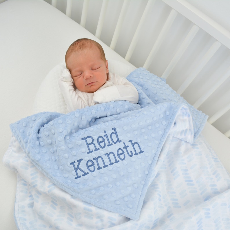May include: A baby sleeps peacefully, swaddled in a light blue blanket. The blanket features a textured, dotted surface and a white patterned underside. Embroidered in blue thread are the names "Reid Kenneth". The scene is set in a white crib.