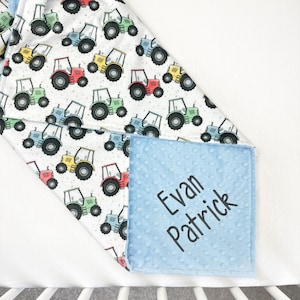 May include: A baby blanket featuring a pattern of colorful tractors in red, green, yellow, and blue on a white background. The blanket has a light blue, textured square with the name "Evan Patrick" printed in black.