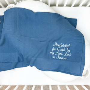 May include: A blue knit baby blanket with the embroidered text "Handpicked for Earth by my Aunt Lexi in Heaven." The blanket is folded over a white pillow in a crib.