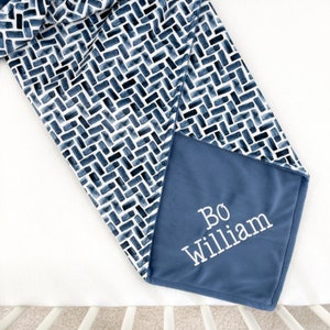 K&ouml;nnte beinhalten: Eine Babydecke mit marineblauem und wei&szlig;em Muster und dem Namen "Bo William" in Wei&szlig; gestickt. Die Decke hat ein geometrisches Ziegeldesign und eine weiche, pl&uuml;schige Textur.