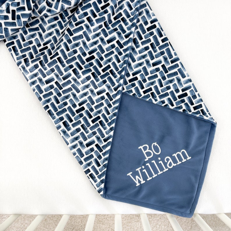 K&ouml;nnte beinhalten: Eine Babydecke mit marineblauem und wei&szlig;em Muster und dem Namen "Bo William" in Wei&szlig; gestickt. Die Decke hat ein geometrisches Ziegeldesign und eine weiche, pl&uuml;schige Textur.