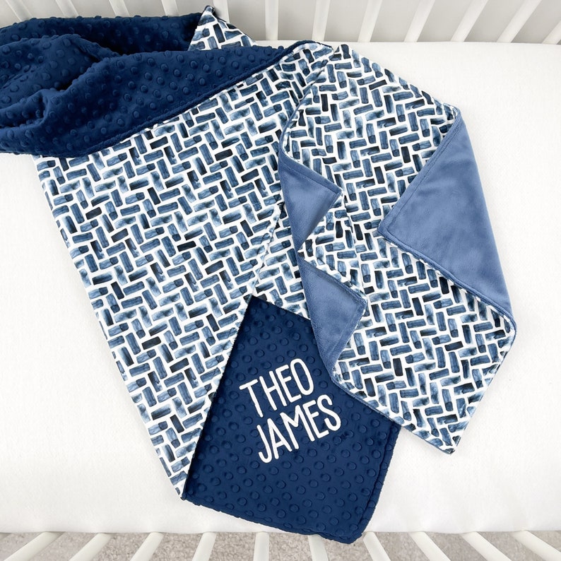 K&ouml;nnte beinhalten: Eine marineblaue Babydecke mit einem geometrischen Aquarellmuster in Blau- und Wei&szlig;t&ouml;nen. Die Decke ist wendbar, mit einer weichen, strukturierten marineblauen R&uuml;ckseite. Der Name "THEO JAMES" ist auf einer kleineren Decke gestickt.