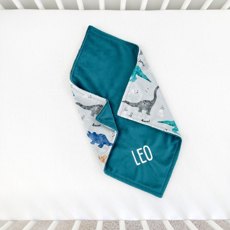 May include: A teal blue baby blanket with a white dinosaur print on one side and the name "LEO" embroidered on the other side.
