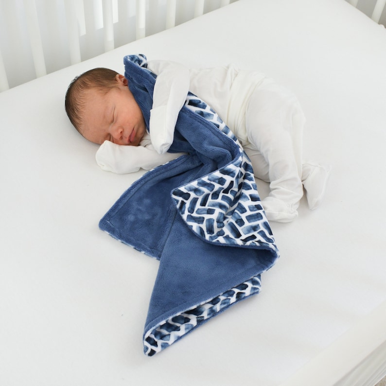 K&ouml;nnte beinhalten: Ein schlafendes Baby liegt in einem wei&szlig;en Kinderbett und ist in eine weiche, blaue Decke geh&uuml;llt. Die Decke hat ein Aquarell-Ziegelmuster in Blau- und Wei&szlig;t&ouml;nen. Die Decke ist aus einem Pl&uuml;schmaterial gefertigt.