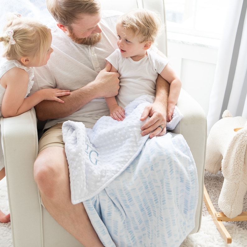 May include: A light blue and white baby blanket with a dotted pattern and a chevron pattern. The blanket has the letter "G" embroidered on it. The blanket is being held by a person.