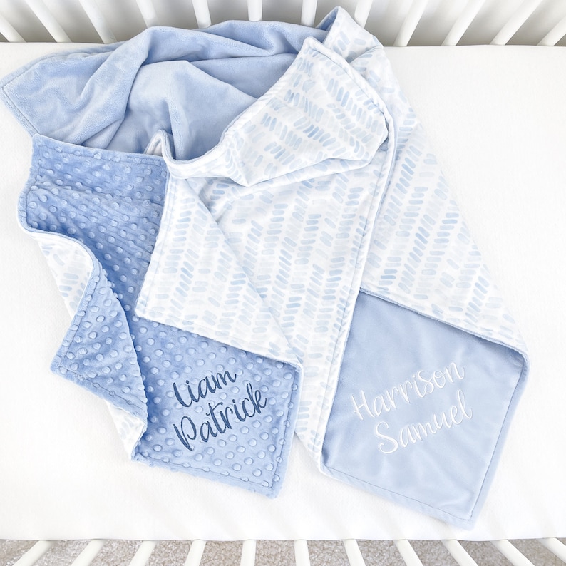 May include: A baby blanket in shades of blue, featuring a soft, textured fabric and a patterned design. The blanket is personalized with the names "Liam Patrick" and "Harrison Samuel" embroidered in elegant script. The blanket is folded on a white surface.