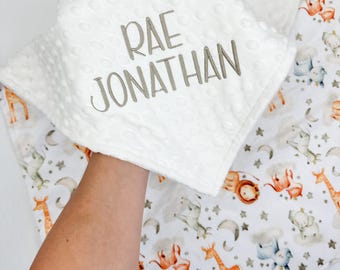 May include: A white baby blanket with a textured, dotted surface, embroidered with the name "RAE JONATHAN" in gray thread. The blanket underneath features a colorful safari animal print, including giraffes, elephants, and lions.
